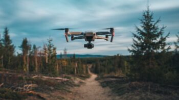 a personal drone flying overhead, equipped with a camera and capturing a photo. The ground below is covered with trees, and there is a trail leading into the forest. The sky is cloudy.