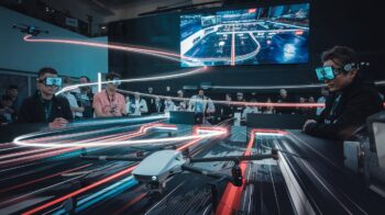 Drone racing event with participants wearing FPV goggles, controlling drones as they navigate a neon-lit track. A large screen in the background displays the course and race progress, with an audience observing the action.